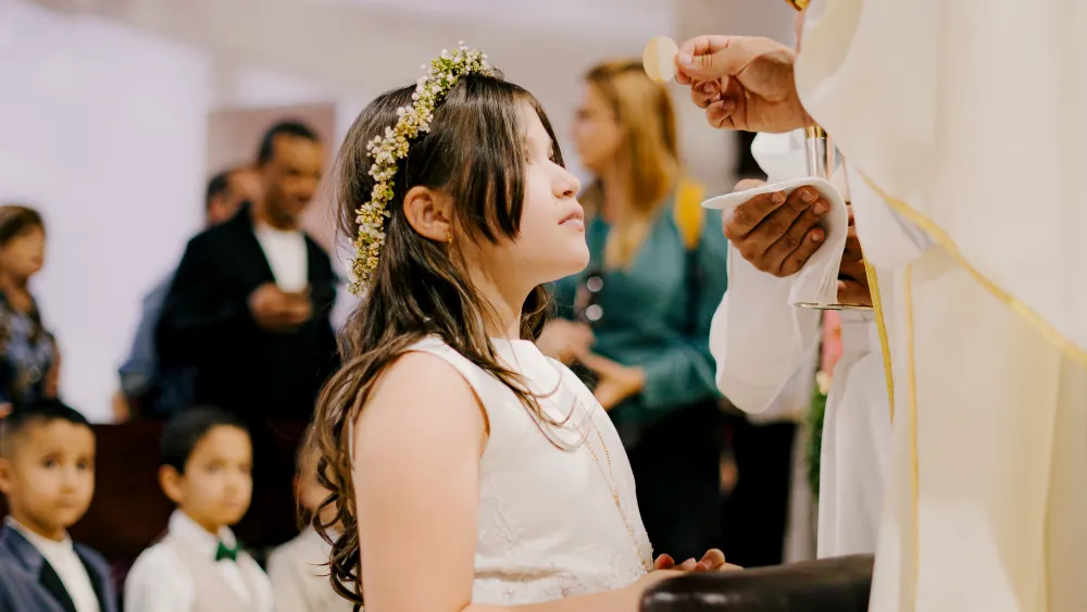 Niña con vestido blanco y corona de flores recibiendo la comunión en la iglesia